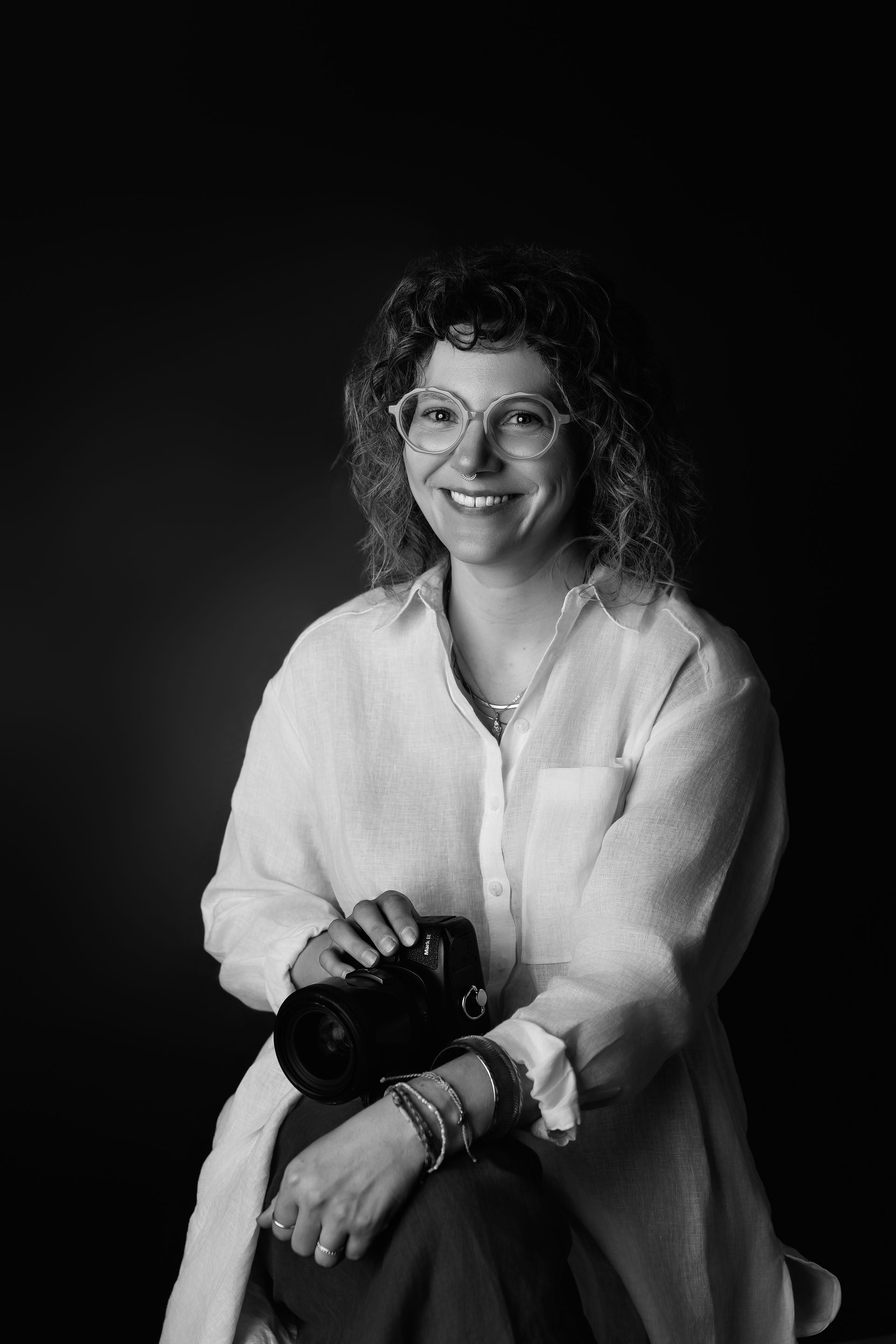 Des Moines, Iowa Headshot and Branding photography - Black and white image of a woman in a white loose fit button up shirt and curly hair and glasses smiles while holding a camera.