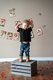 Des Moines, Iowa children and family photographer - toddler boy with curly blonde hair stands by a wooden crate and flexes his muscles. Valentine's Day decor and red rose petals surround him.