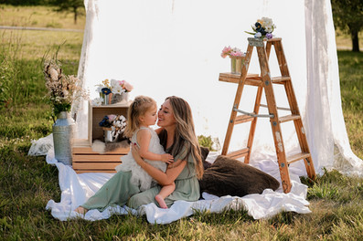 Des Moines, Iowa Family Photographer - a mother and her toddler-aged daughter are sitting outside in front of a white lace curtain . Mom is holding the child in her lap and looking at her.