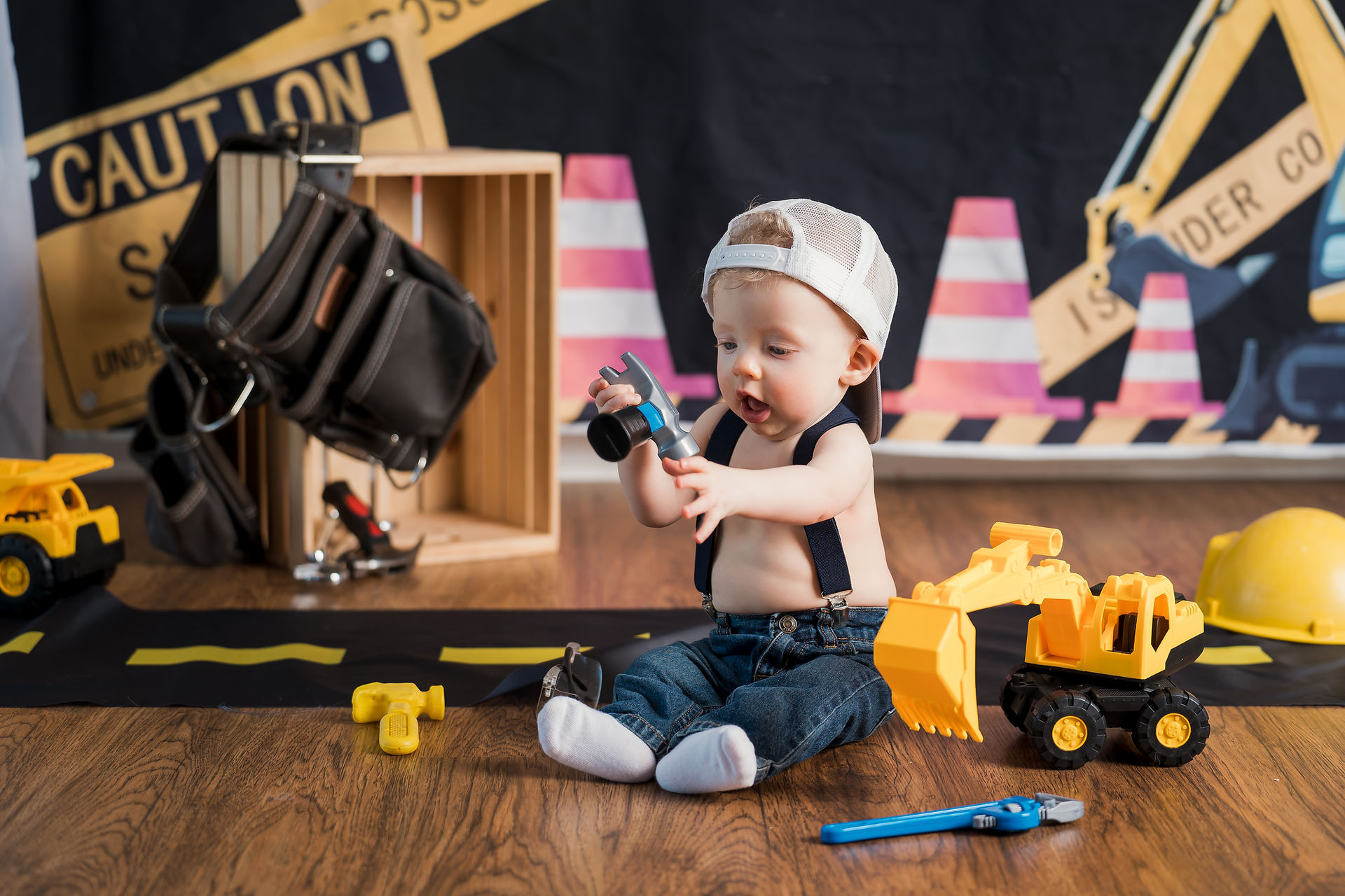Des Moines, Iowa photographer - infant boy is sitting in studio for a constuction themed photo session. He is wearing denim jeans, suspenders, and backwards cap and is holding a hammer.