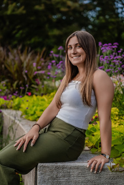 Des Moines, Iowa senior photography - dark haired teenager girl in a white tank top poses on a ledge with purple and green florals behind her.