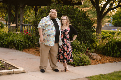Norwalk and Des Moines, Iowa family photographer - adult woman in a long floral dress poses with her adult son in a black with Hawaiian shirt over top, and khaki pants in a wooded area during sunset.