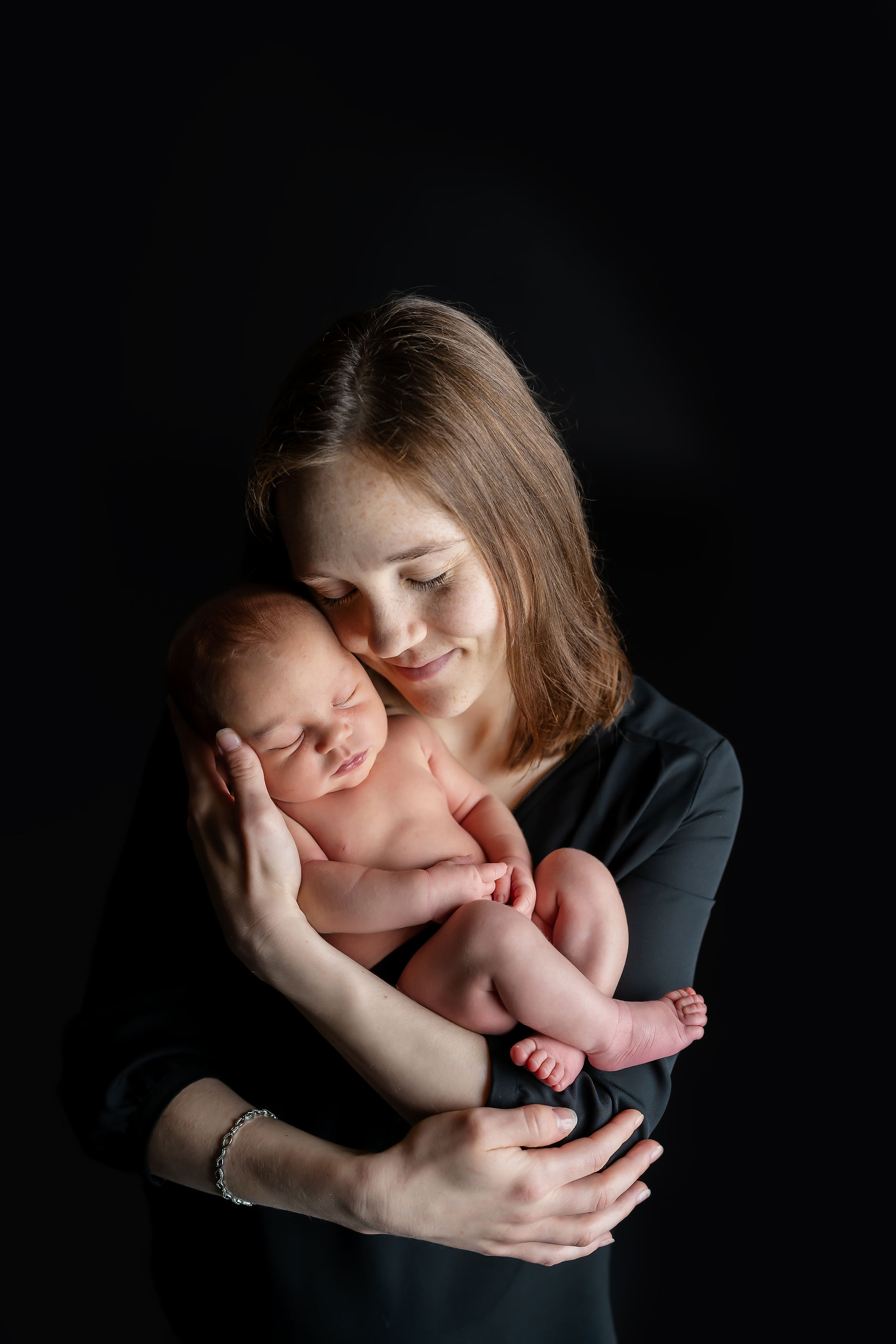 Des Moines, Iowa Newborn Photographer - A new mother wearing a black shirt is posed cheek to cheek with her newborn baby girl with their eyes closed.