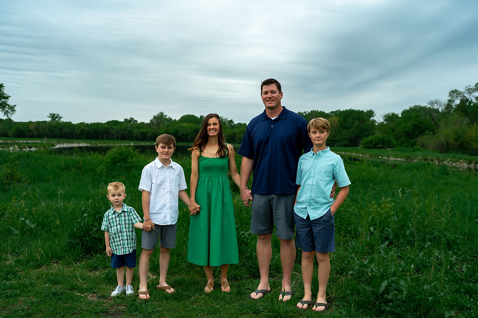 Des Moines, Iowa Family Photographer - A family of five (Mom, Dad, and three boys) holding hands in a green field with a pond in the background.
