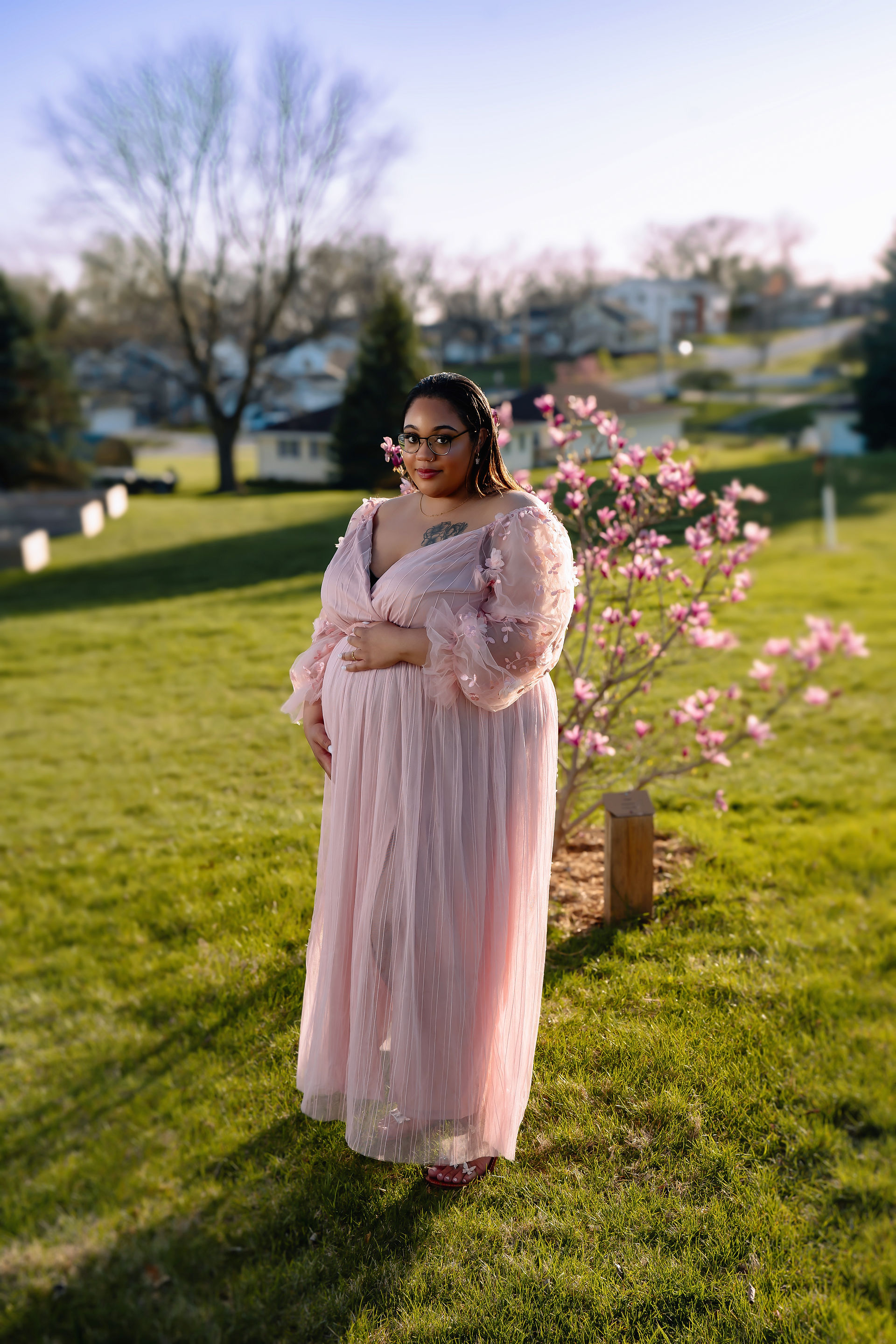 Des Moines family and maternity photographer - pregnant black female in a pink gown holds her belly in front of a pink magnolia tree.