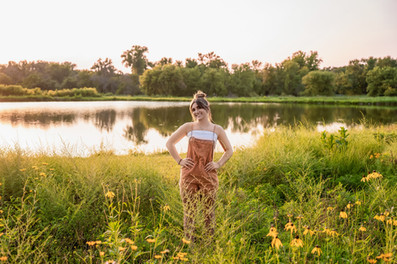 Des Moines, Iowa senior photography - a girl with dark hair styled in a messy bun wearing a white tank top and orange short overalls is standing in a field of yellow flowers with a pond in the background at sunset.