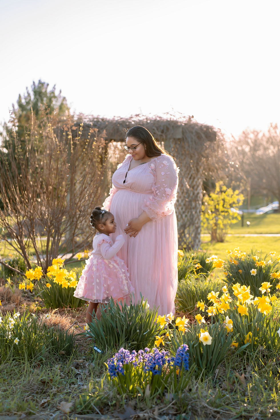 Des Moines - family and maternity photographer - 36 week pregnant black mother hugs her toddler daughter surrounded by flowers-both wearing pink dresses.