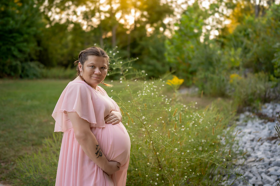 Norwalk and Ames, Iowa maternity and newborn photographer - a pregnant woman in a light pink gown is standing in front of white flowers at sunset holding her belly.