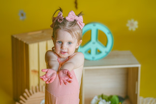 Calli Tuggle Photography - Des Moines Family Photographer - Toddler girl in pink bell-bottom jumpsuit and hair bows is in studio with yellow walls and floor decorated in 1960's themed set reaching towards camera
