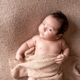 Des Moines, Iowa Newborn Photography - newborn baby boy with dark hair on a tan background with tan swaddle over him looking up and smiling