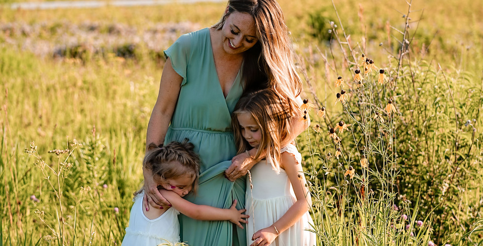 Des Moines, Iowa Family Photographer - a mother and her two daughters are hugging while standing in a prairie during golden hour.
