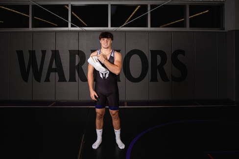 Norwalk and Des Moines, Iowa high school senior photography - high school wrestler carries his wrestling shoes over his shoulder with his school logo in the background of the wrestling room.