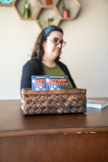 Des Moines, Iowa Branding Photographer - author Stephanie R. Caffrey poses sitting at a desk with a stack of her debut novel "Mistaken Identify"