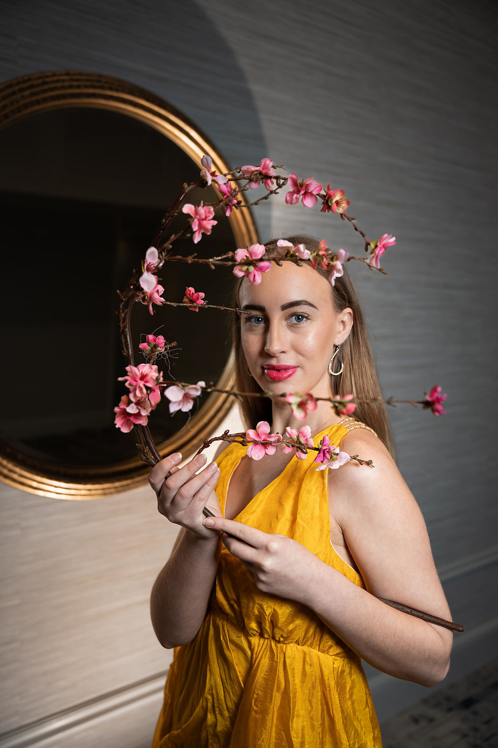 Des Moines, Iowa portrait photographer - young adult woman is wearing a yellow gown and silver hoop earrings, holding pink orchids fframing her face, an orate gold mirror is in the background.
