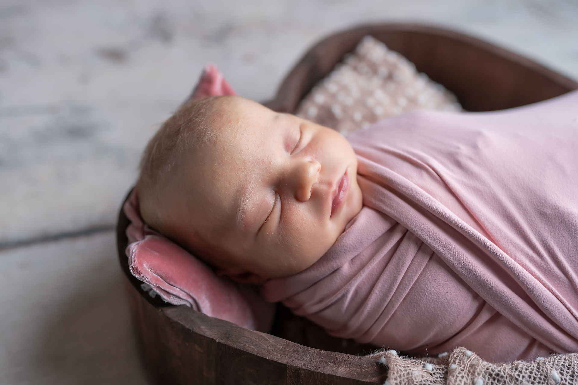 Des Moines, Iowa Newborn Photographer - infant baby girl is wrapped in a pink swaddle and is lying in a wooden heart bowl on a wooden floor--close up of facial features.
