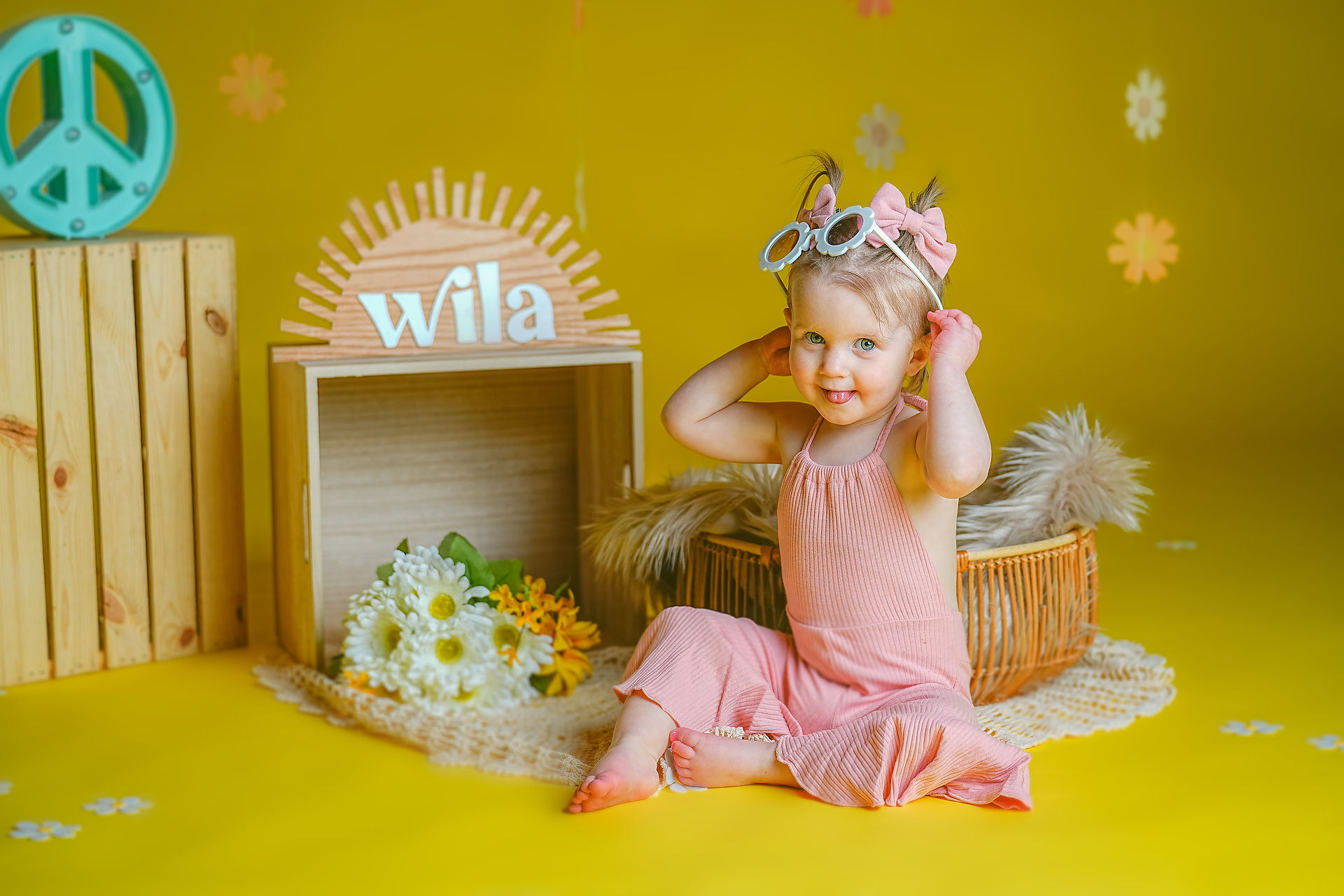 Calli Tuggle Photography - Des Moines Family Photographer - Toddler girl in pink bell-bottom jumpsuit and hair bows is in studio with yellow walls and floor decorated in 1960's themed set trying on white sunglasses and smiling at camera
