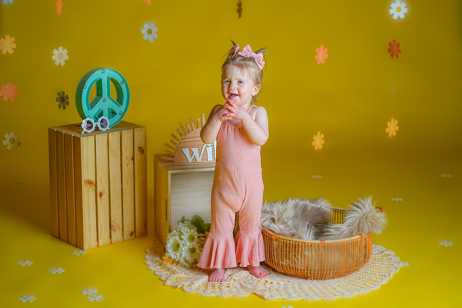 Calli Tuggle Photography - Des Moines Family Photographer - Toddler girl in pink bell-bottom jumpsuit and hair bows is in studio with yellow walls and floor decorated in 1960's themed set holding an orange pacifier smiling big