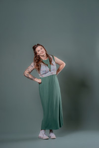 Des Moines and Norwalk, Iowa photography - adult woman with long hair is wearing an ankle-length green dress and a floral denim vest is standing with her hands on her hips in front of a green backdrop.