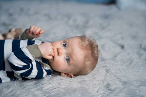 Des Moines, Iowa photographer - infant boy with blonde hair and blue eyes is lying on a gray fur rug with a lightblue backdrop. He is wearing a navy and white striped outfit and smiling.