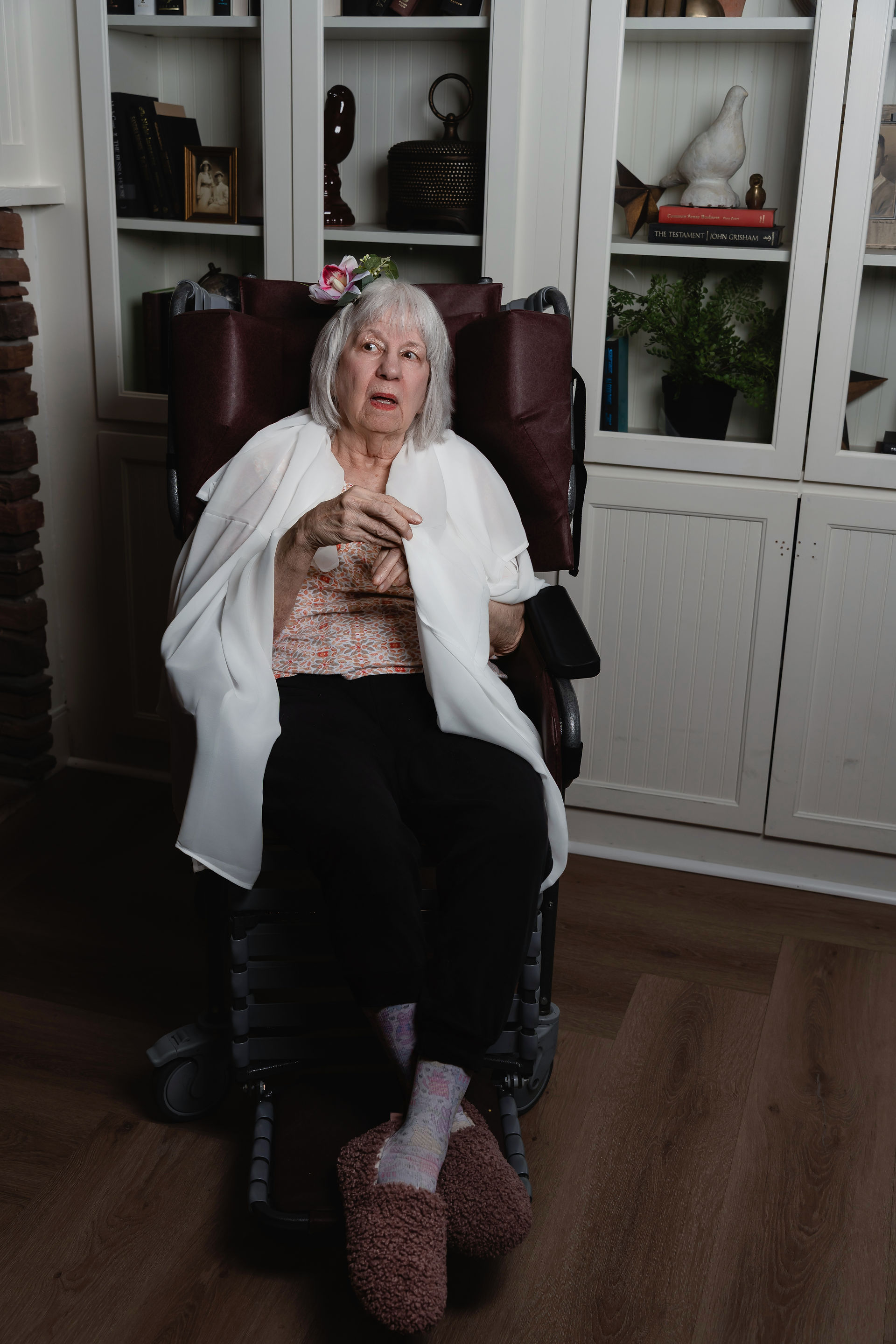 Des Moines, Iowa portrait photographer Calli Tuggle Photography - elderly adult woman in a wheelchair wears a sheer white blouse over a pink blouse with a pink flower in her hair against a white bookshelf.