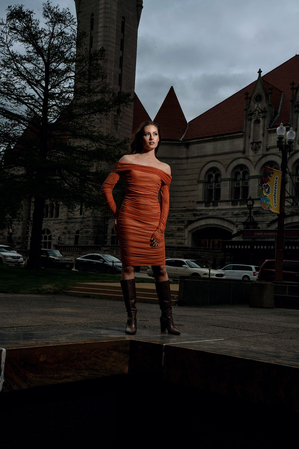 Des Moines, Iowa - women's portrait photographer - young adult woman with dark hair is posed outdoors on an overcast day wearing an off the shoulder orange dress, looking away from the camera.