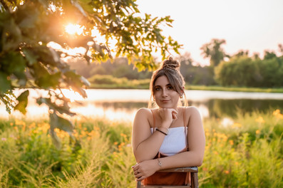 Des Moines, Iowa senior photography - a girl with dark hair styled in a messy bun wearing a white tank top and orange short overalls is leaning against a wooden ladder during sunset.