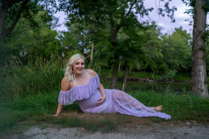 Des Moines and Norwalk, Iowa family photographer - pregnant blonde female wearing a light purple gown is posed sitting in the grass on front of tall grasses, trees, and the Des Moines River. She is looking at the camera, smiling, and holding her belly.
