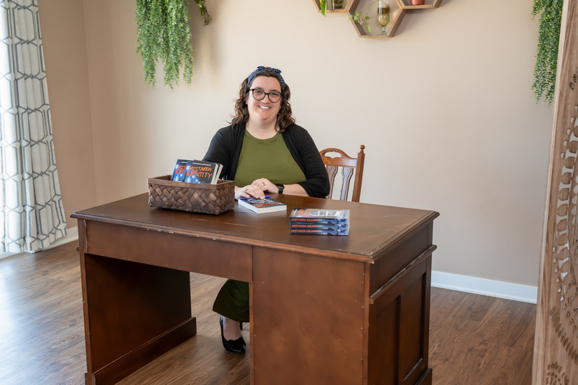 Des Moines, Iowa Branding Photographer - author Stephanie R. Caffrey poses sitting at a desk with a stack of her debut novel "Mistaken Identify"