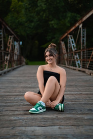 Des Moines, Iowa senior photographer - teenager girl with dark hair pulled into a messy bun is sitting on a wooden and metal bridge.