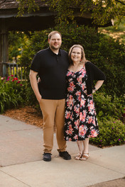 Norwalk and Des Moines, Iowa family photographer - adult woman in a long floral dress poses with her adult son in a black polo and khaki pants in a wooded area at sunset.