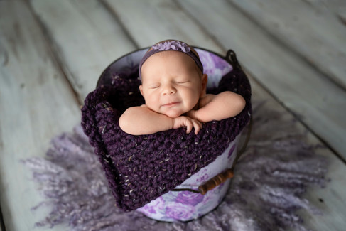 Des Moines and Norwalk, Iowa newborn photographer - a baby girl sits in a purple bucket for newborn photos and smiles