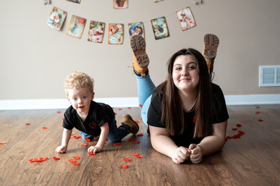 Des Moines, Iowa teen and children's photography - teen girl with long dark hair is lying on a wooden floor surrounded by red rose petals while toddler brother explores the roses.