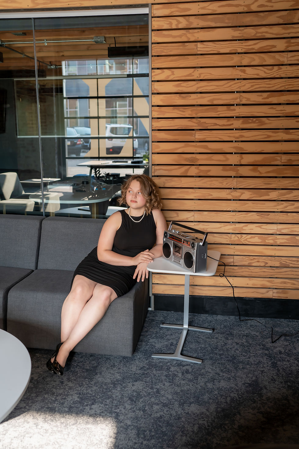 Des Moines Senior Photographer - Teen/Young adult woman in a black dress in high heels is sitting on a gray couch with a wood paneled wall behind her, looking off into the distance.