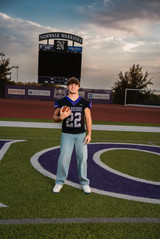 Norwalk and Des Moines, Iowa high school senior photography - football player stands on the field wearing his jersey and jeans holding a football in front of an unlit scoreboard during golden hour.