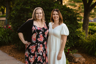 Norwalk and Des Moines, Iowa family photographer - adult woman in a long floral dress poses with her adult daughter in a white dress in a wooded setting at sunset.