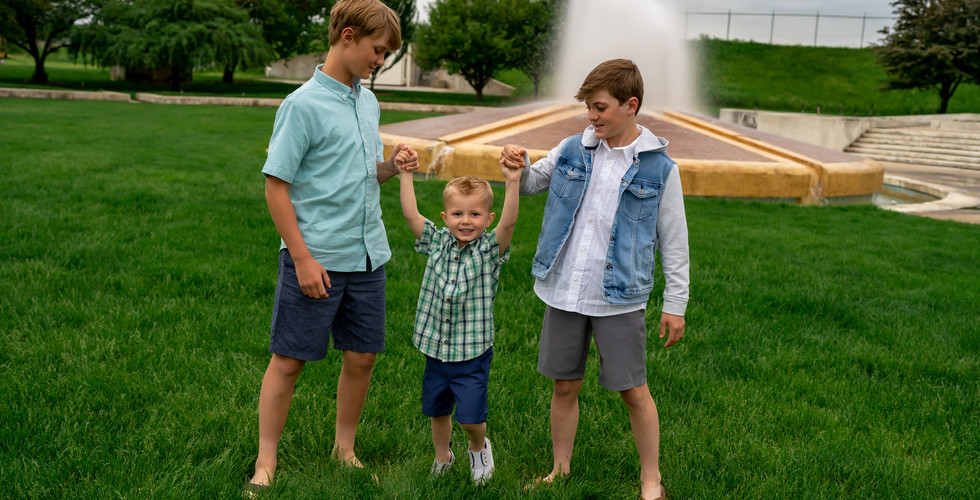 Des Moines, Iowa Family Photographer - Two boys are holding their toddler brother's hand and swinging him between them. There's green grass and a fountain in the background.
