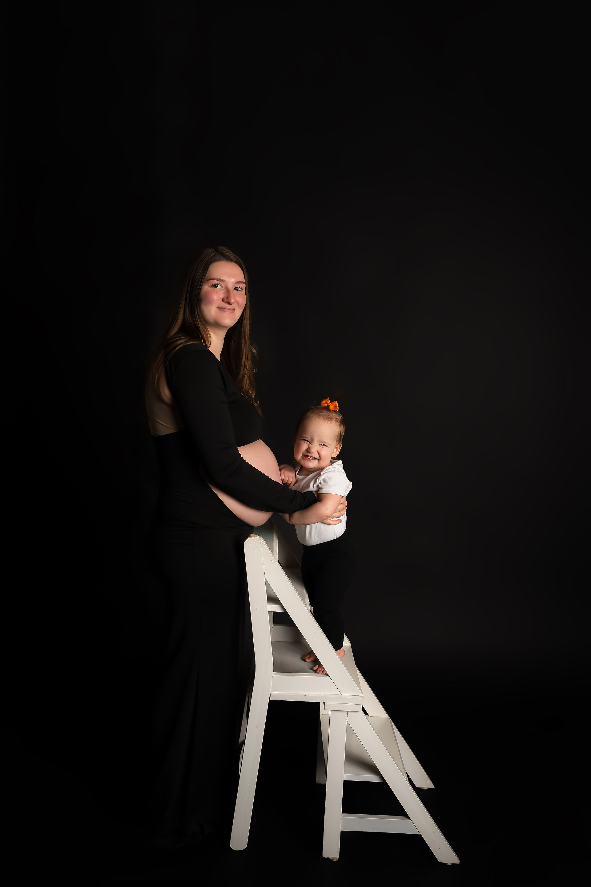 Des Moines and Norwalk, Iowa pregnancy photos - mother in black maternity gown with exposed belly stands next to toddler toddler on white ladder, both looking at the camera and smile.