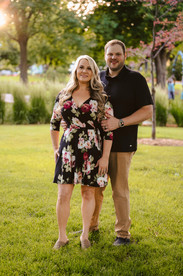 Norwalk and Des Moines, Iowa familyw photographer - adult woman with long blonde hair and a short floral dress poses with her husband in a black polo and khaki pants in a wooded park during sunset.