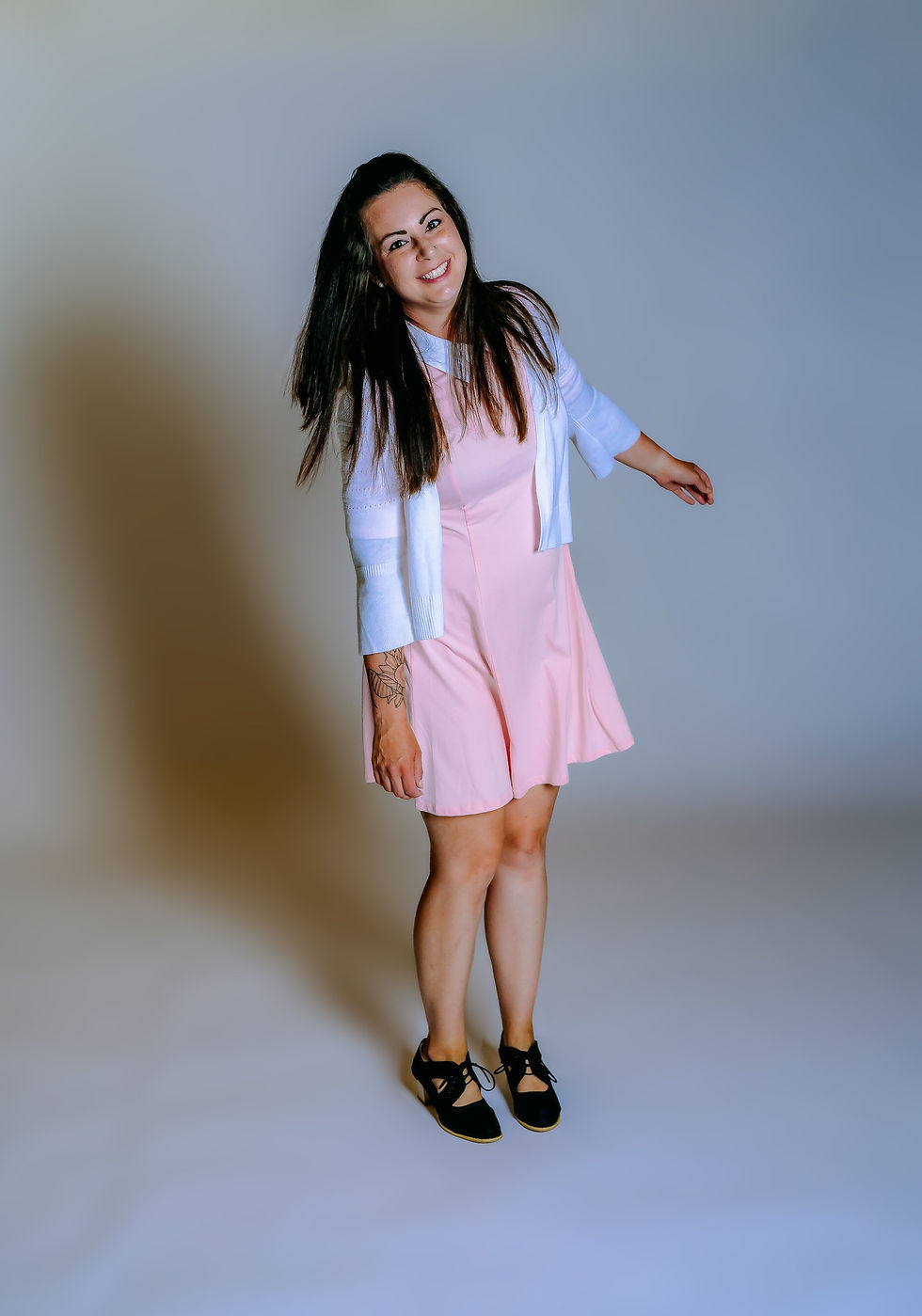 Des Moines Photographer - dark haired woman is posing in studio on a white backdrop. She is wearing a light pink dress with a white collar, a white cardigan sweater, and black oxford shoes.