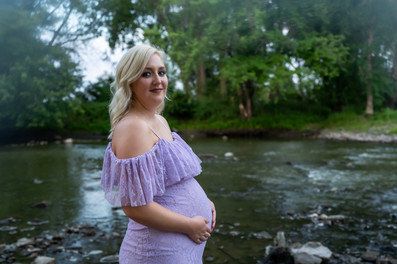 Des Moines and Norwalk, Iowa family photographer - pregnant blonde female wearing a light purple gown is posed standing in front of the Des Moines River looking at the camera and holding her belly.
