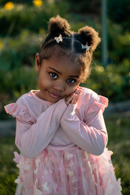 Des Moines family and children's photographer - black female toddler wearing a pink dress and butterfly clips in her hair poses with hands by her face and a sly smile.