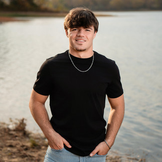 Des Moines and Norwalk, Iowa high school senior photographer - male teenager in a black t-shirt and jeans stands in front of a lake smiling with his hands in his pockets.