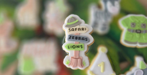 Safari themed decorated cookie shaped like a wooden signpost. Four arrows point to giraffe, zebra, tiger and elephant illustrations. 