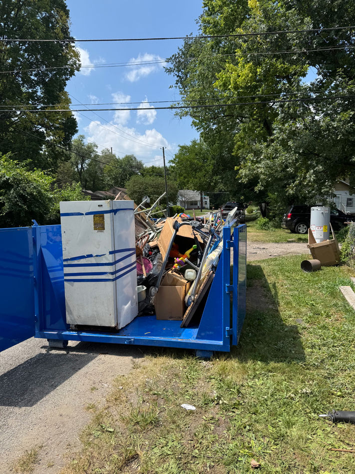 A blue dumpster sits on the side of the road. The back end of the dumpster is wide open and there is a fridge and other junk items inside.