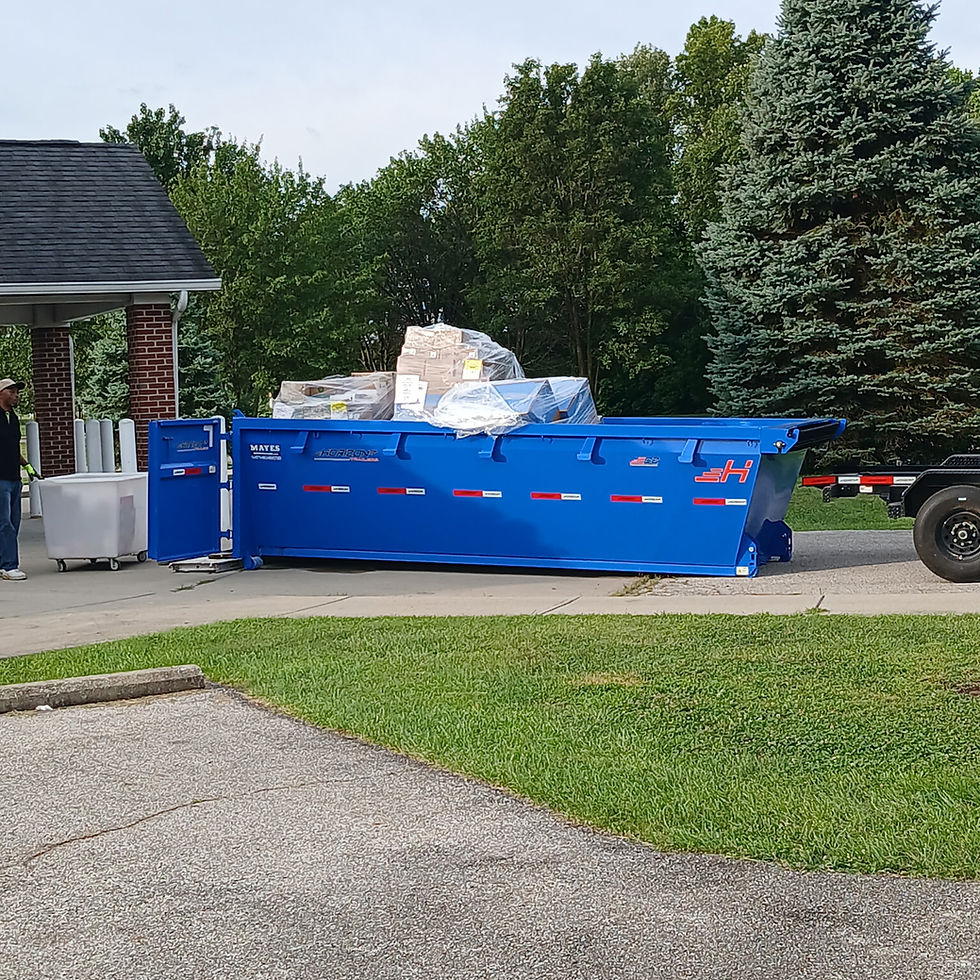 A blue dumpster filled with boxes wrapped in plastic wrap, sits behind a black trailer, it's back double doors are open and a person has roleld a white cart up to the trailer.