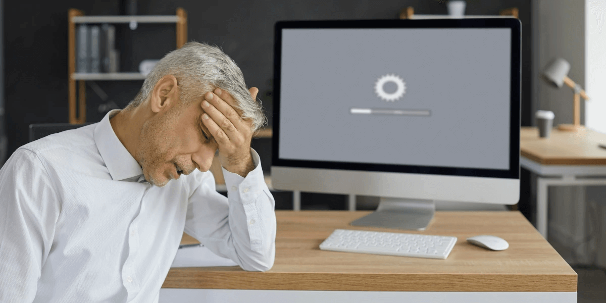 An office worker sitting with hand to forehead and in front of a computer, waiting for a slow site to fully load the page