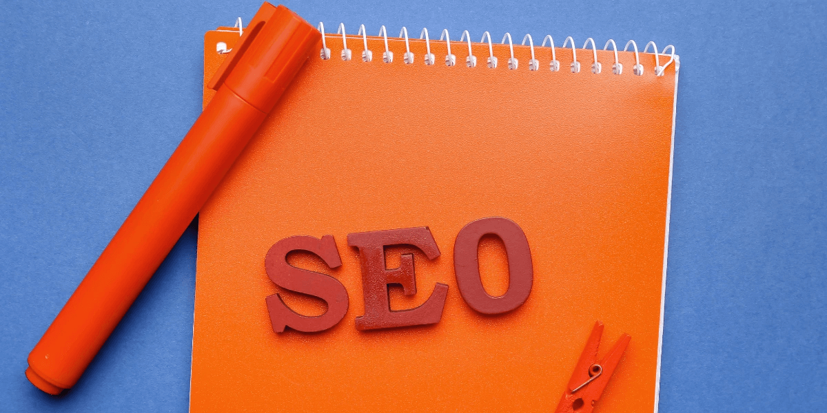 An orange notebook against a blue background with a pen and letter cutouts forming the word SEO on top