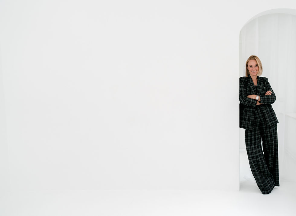 Carolin steht lächelnd im karierten Anzug mit verschränkten Armen vor einer weißen Wand.