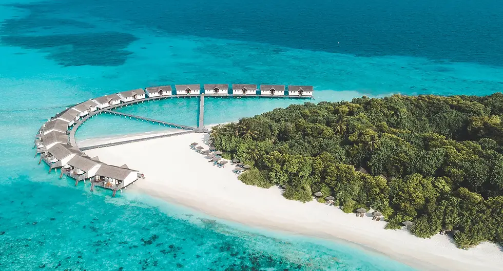 Aerial view of a tropical island with white sandy beaches, lush greenery, and a curved pier stretching into turquoise waters. Overwater bungalows line the pier, surrounded by the clear blue ocean, creating a serene and luxurious resort setting.