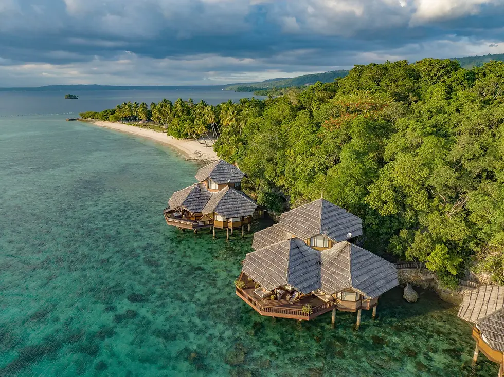 Aerial view of a tropical beach resort with overwater bungalows built on stilts above clear turquoise water. The white sandy beach is lined with palm trees and lush greenery, with a distant island visible on the horizon. The scene captures a serene and luxurious vacation setting.